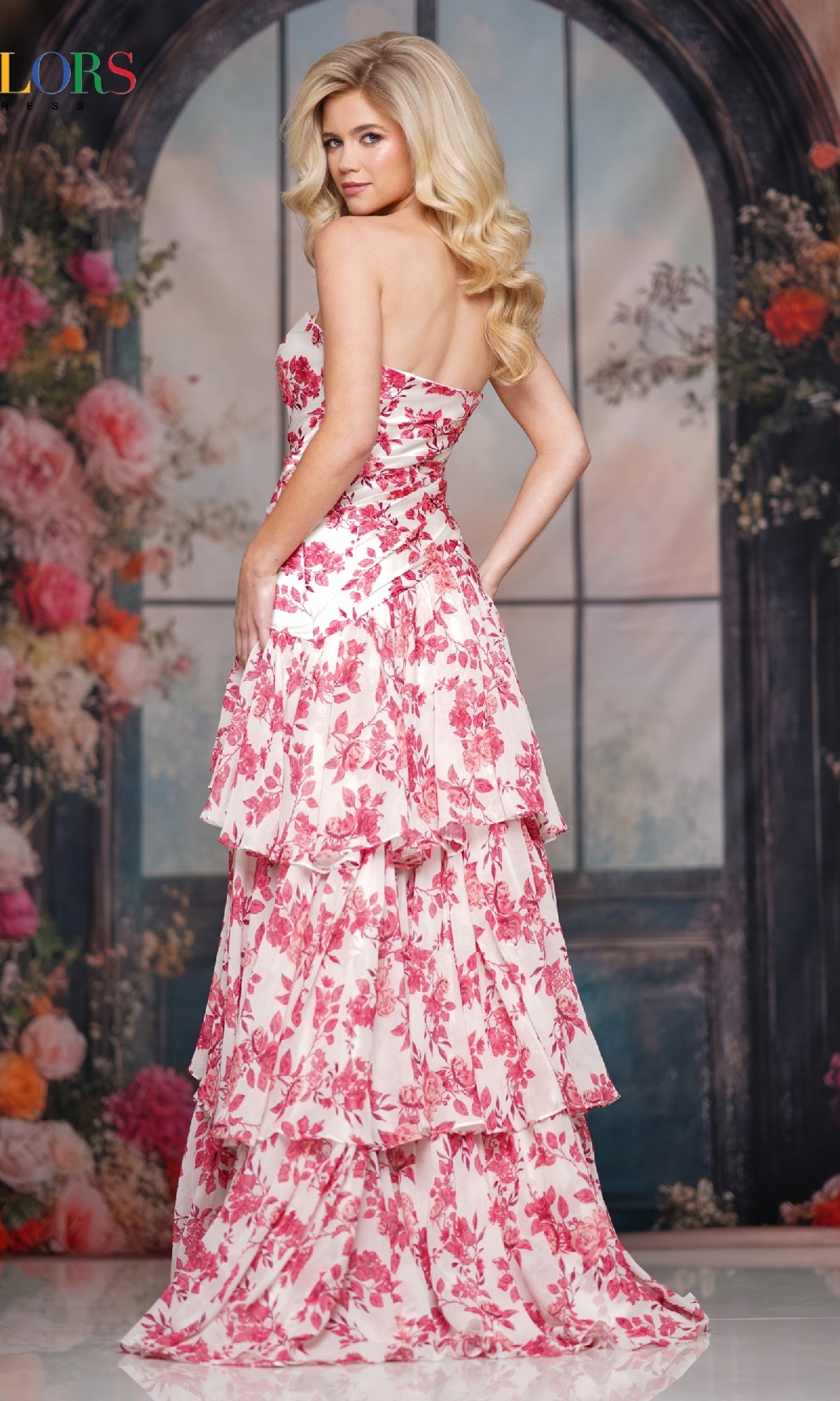 A woman in the Strapless Long Print Prom Dress: Colors Dress 3888, featuring pink and white tiers, stands before a window backdrop with flowers and greenery.